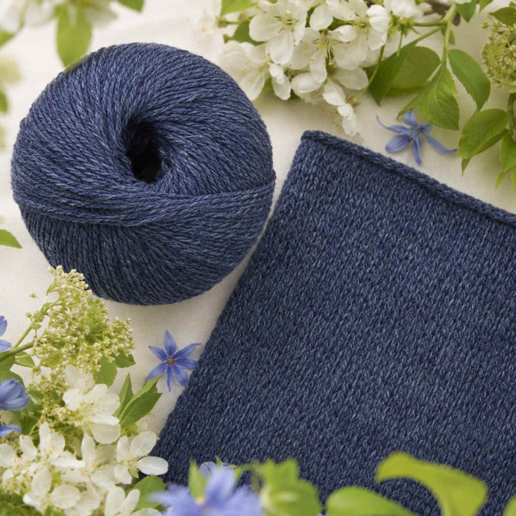 Mørkere blå garn uden banderole vist som garnnøgle og strikket prøve med tydelig, blød merinostruktur, omgivet af hvide blomster, lilla blomster og grønne blade på et rustikt træbord.