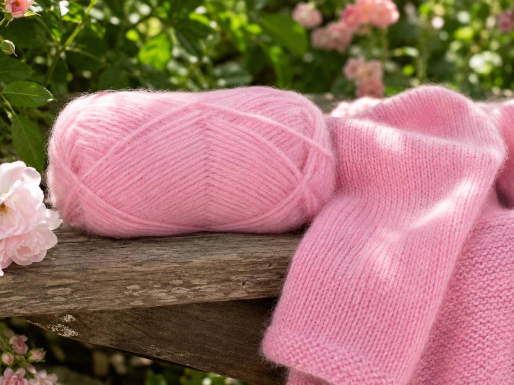 ALT tekst: garnnøgle uden banderole i lys rosa med matchende blødt og tæt strikket blow yarn-projekt på en rustik havebænk i en blomstrende have.