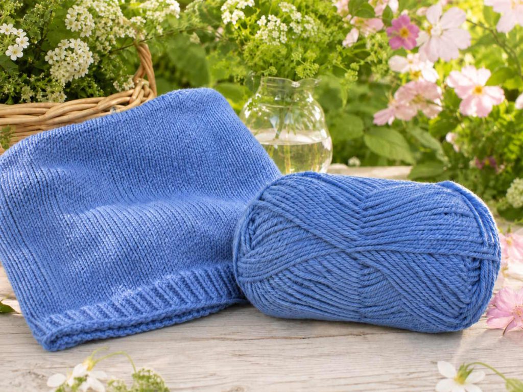 Det blå garn ligger på et havebord der er omgivet af blomster - der er ved at blive strikket en bluse til et barn