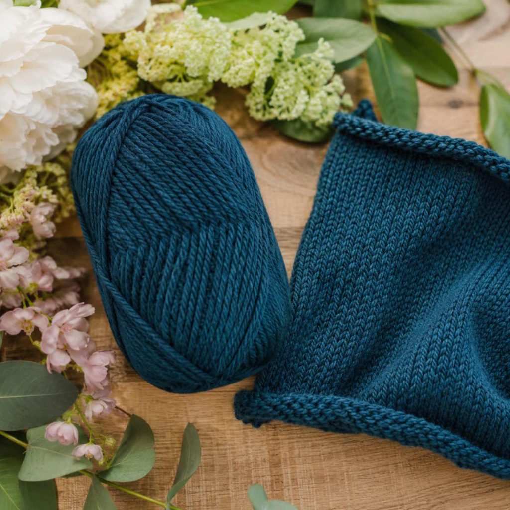 Blåt garn uden banderole vist som garnnøgle og strikket prøve med tydelig uldstruktur, omgivet af blomster og grønne blade på et træbord.