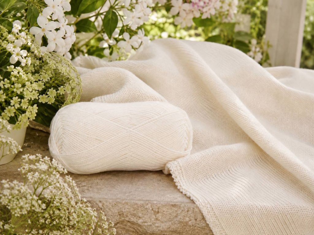 Hvidt garn med strikket stykke ligger på en stol i haven omgivet af blomster og grønt